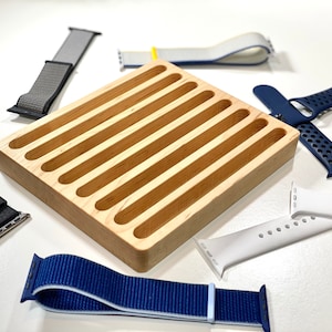 May include: A wooden watch band organizer with eight slots. The organizer is made of light wood and has a smooth finish. There are six watch bands around the organizer, including a black leather band, a gray nylon band, a white silicone band, a blue nylon band, a white and blue nylon band, and a blue and black sport band.
