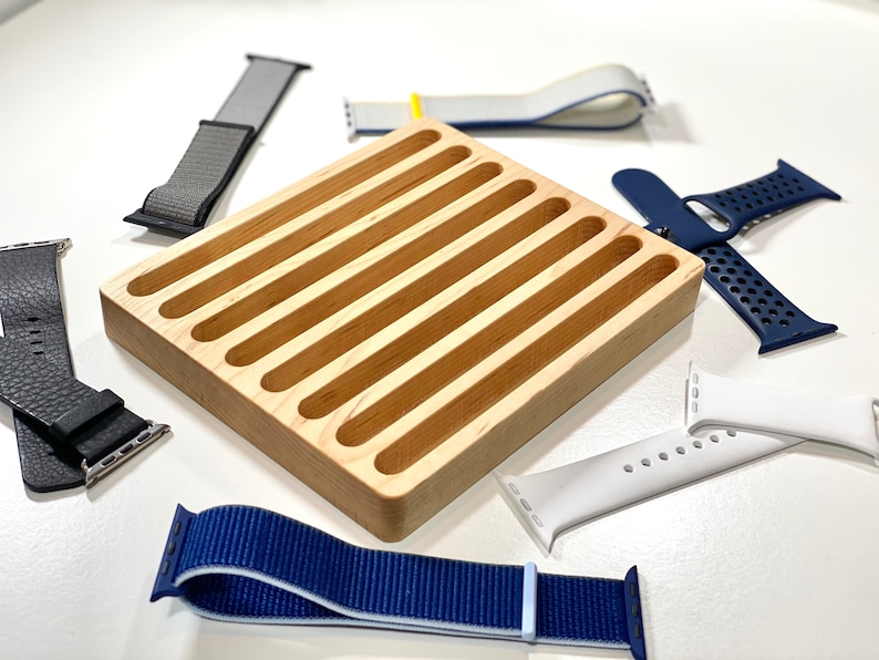 May include: A wooden watch band organizer with eight slots. The organizer is made of light wood and has a smooth finish. There are six watch bands around the organizer, including a black leather band, a gray nylon band, a white silicone band, a blue nylon band, a white and blue nylon band, and a blue and black sport band.