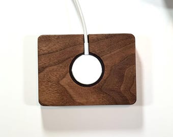 May include: A wooden charging stand for an Apple Watch, featuring a circular cutout for the watch face and a slot for the charging cable. The stand is made of dark brown walnut wood.