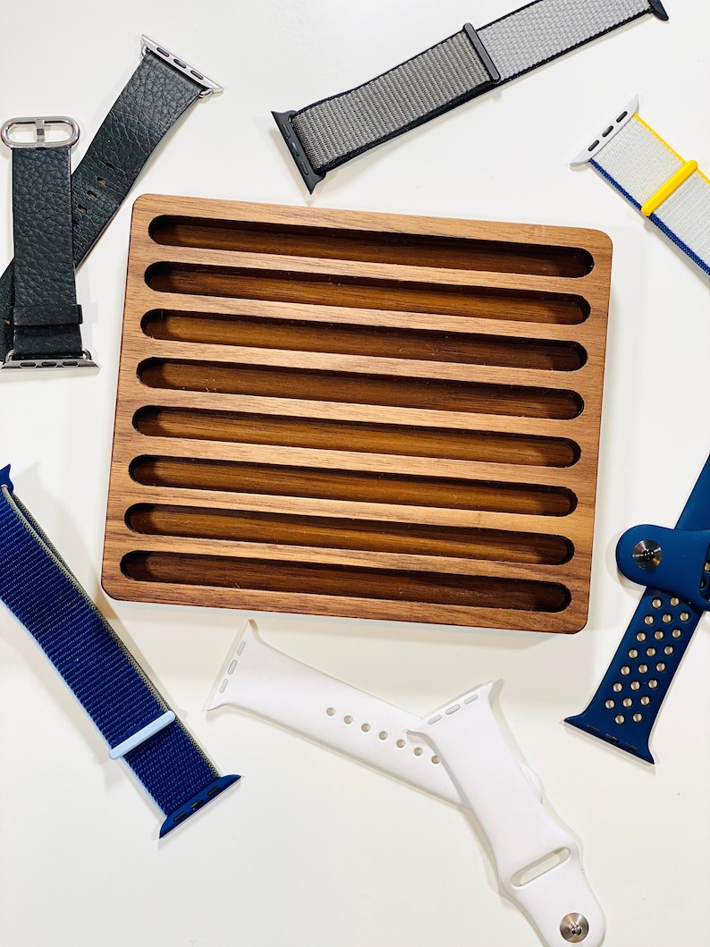 May include: A wooden watch band organizer with eight slots for storing watch bands. The organizer is made of light brown wood and has a smooth finish. There are several watch bands in the image, including a black leather band, a gray nylon band, a white silicone band, a blue nylon band, and a white and yellow nylon band.