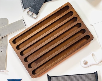 May include: A wooden watch band organizer with six slots for storing watch bands. The organizer is made of dark brown wood and has a smooth finish. There are several watch bands in the image, including black leather, gray nylon, and blue silicone.