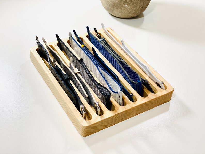 May include: A wooden watch band organizer with eight slots to hold different watch bands. The organizer is made of light brown wood and has a natural finish. The watch bands are a variety of colors, including black, gray, blue, and white.