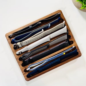 May include: A wooden watch band organizer with six slots, holding a variety of watch bands in different colors and materials, including black, white, blue, and gray.