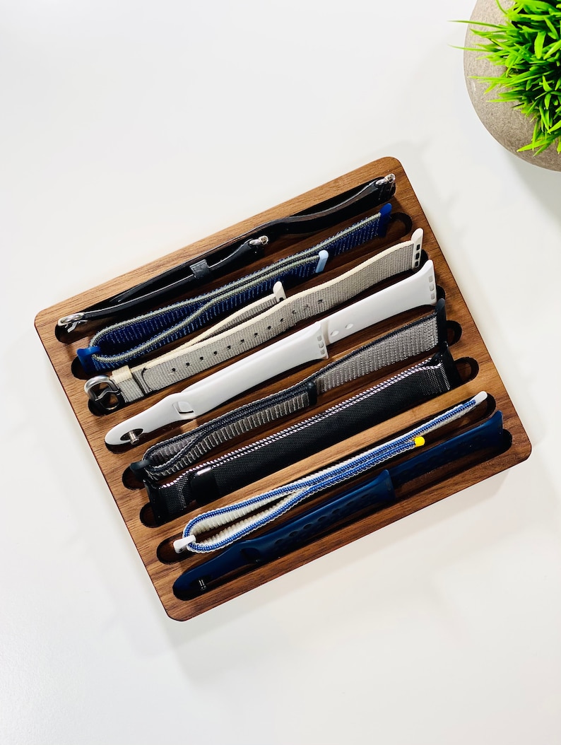May include: A wooden watch band organizer with six slots, holding a variety of watch bands in different colors and materials, including black, white, blue, and gray.
