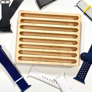 May include: A wooden watch band organizer with eight slots for storing watch bands. The organizer is made of light brown wood and has a smooth finish. There are several watch bands in the image, including a black leather band, a gray nylon band, a white silicone band, a blue nylon band, and a yellow and white nylon band.