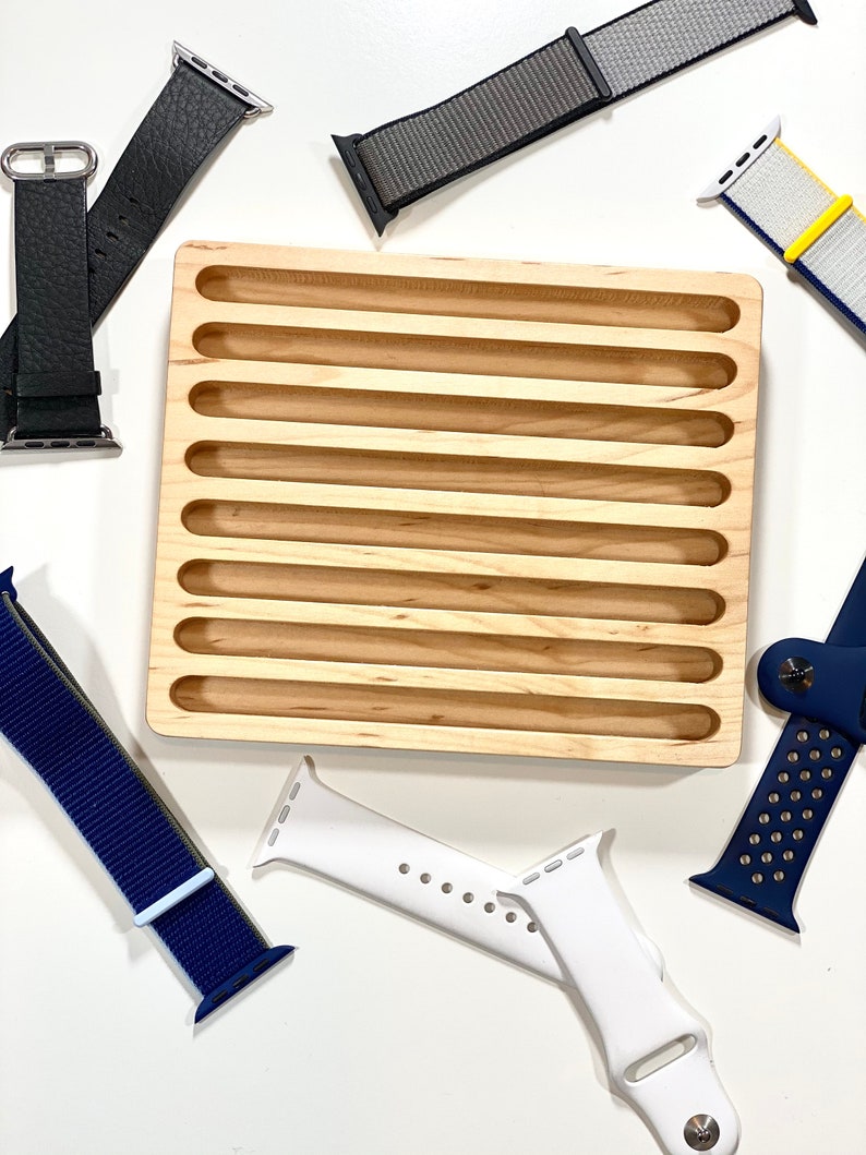 May include: A wooden watch band organizer with eight slots for storing watch bands. The organizer is made of light brown wood and has a smooth finish. There are several watch bands in the image, including a black leather band, a gray nylon band, a white silicone band, a blue nylon band, and a yellow and white nylon band.