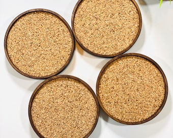May include: Four round cork coasters with a dark brown wood frame. The coasters are arranged in a square shape on a white surface.