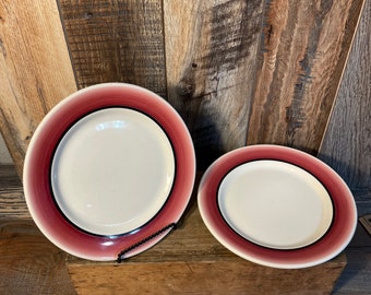 May include: Two ceramic plates with a white center and a red and black rim. One plate is round, and the other is oval. The plates are displayed on a wooden surface against a wooden background.