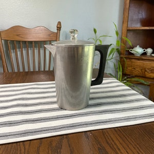 May include: A silver coffee pot with a black handle and a clear glass lid. The pot is sitting on a white and black striped placemat.
