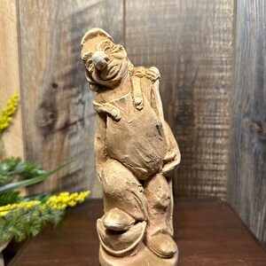 May include: A whimsical ceramic figurine of a person wearing overalls and a hat, holding a bucket. The figurine is painted in a light brown color and has a textured surface.