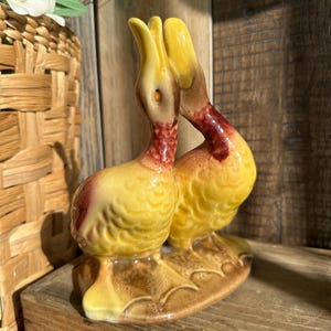 May include: A ceramic figurine of two ducks, facing each other. The ducks have yellow bodies, red necks, and brown accents. They are perched on a brown base. The figurine is a decorative item.