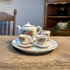 May include: A vintage white porcelain tea set with pink rose floral designs. The set includes a teapot, sugar bowl, two teacups, and saucers, all arranged on a round serving plate. The tea set is placed on a wooden table.