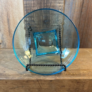 May include: A clear, turquoise glass bowl with a square center and a round outer rim. The bowl is supported by a black metal stand. The bowl is displayed on a wooden surface.