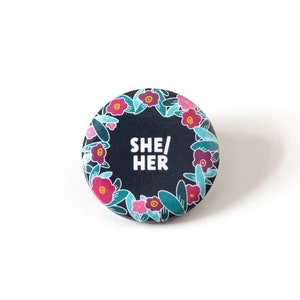 Könnte beinhalten: Ein runder Button mit dunkelblauem Hintergrund und dem weißen Text "SHE/HER". Der Button ist von einem floralen Kranz mit rosa und lila Blumen und grünen Blättern umgeben.