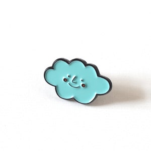 May include: A light blue enamel pin shaped like a cloud with a smiling face. The cloud has black outlines and brown eyes.