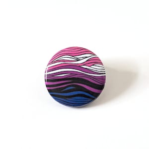 May include: A round button with a wavy design in pink, white, blue, and black colours.
