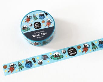 May include: Two rolls of washi tape with a light blue background and a repeating pattern of cartoon cats in space suits, planets, and spaceships. One roll is unrolled, showing the tape's design. The text "Give me space" is visible.