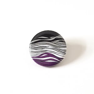 May include: A round button featuring a black, white and purple wavy pattern. The button represents the asexual pride flag.