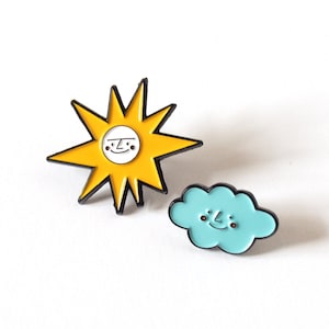 Może przedstawiać: Dwie szpilki emaliowane, jedna przedstawiająca żółte słońce z uśmiechniętą twarzą, a druga niebieski obłok z uśmiechniętą twarzą.