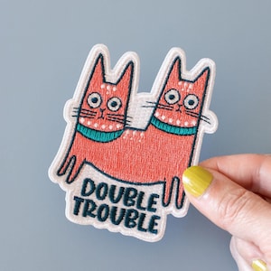 May include: Embroidered patch featuring two orange cats joined together, with teal collars and wide eyes. The patch has the words "DOUBLE TROUBLE" in teal lettering. The patch is held against a light blue background.