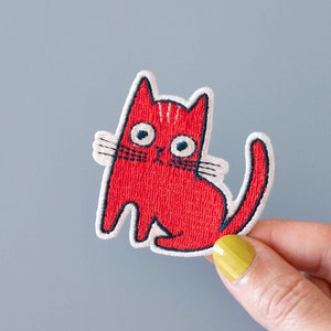 May include: Red embroidered iron-on patch of a cat with white whiskers and blue eyes.