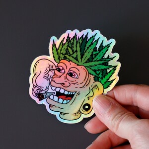 Peut inclure: Autocollant holographique repr&eacute;sentant un visage de dessin anim&eacute; avec des feuilles de cannabis vertes en guise de cheveux, un joint allum&eacute; et un large sourire. L'autocollant a un &eacute;clat arc-en-ciel et une boucle d'oreille noire.