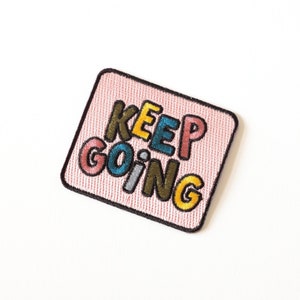 Peut inclure: Un patch en tissu rose avec le texte "KEEP GOING" en lettres brodées colorées.