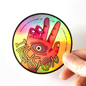 May include: A round, holographic sticker featuring a hand with an eye in the palm, making a peace sign. The word "ILLUSION" is written in a stylized font. The sticker has a rainbow gradient background.