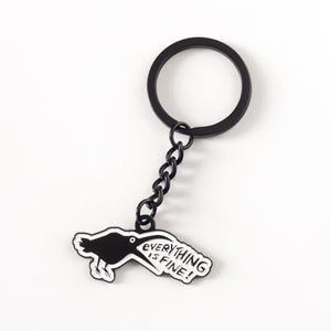 May include: A white ceramic mug with a black handle and the text "But First, Coffee" in a bold, black font. The mug is set against a blurred background. The mug appears to be approximately 10 cm tall.