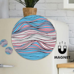 May include: A round magnet with a blue, pink, and white wavy pattern, resembling the transgender pride flag. The word "MAGNET" is printed below a graphic of a horseshoe magnet. Two smaller magnets with a similar design are also visible.
