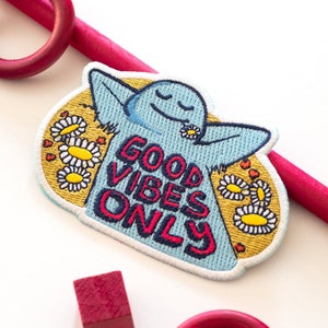 K&ouml;nnte beinhalten: Ein blauer gestickter Patch mit einer Comicfigur mit geschlossenen Augen und einer G&auml;nsebl&uuml;mchen in den Haaren. Der Patch hat gelbe G&auml;nsebl&uuml;mchen und den Text "GOOD VIBES ONLY" in Rot und Blau.