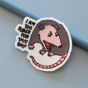 Op de afbeelding: Gestikte patch met een cartoon-opossum met een grijnzende bek en de tekst "I am too sober for this shit".