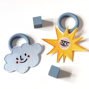 Może przedstawiać: Dwa haftowane naszywki, jedna to niebieska chmura z uśmiechającą się twarzą, a druga to żółte słońce z uśmiechającą się twarzą. Obie naszywki mają czerwone policzki.