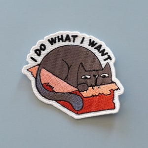 May include: A white embroidered patch with a black cat lying in a red and orange box. The text "I do what I want" is embroidered in black around the cat.