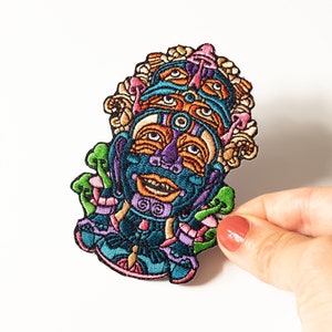 Może przedstawiać: Kolorowa haftowana łatka przedstawiająca postać z wieloma oczami i twarzami o psychodelicznym wzorze. Postać otoczona jest grzybami i innymi elementami florystycznymi.