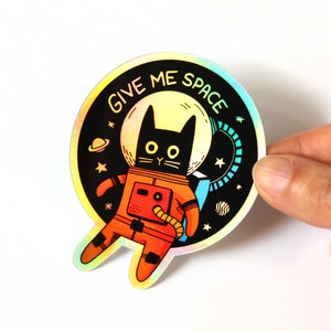 May include: A holographic sticker featuring a black cat wearing a red space suit with the text "Give Me Space" in a rainbow font.