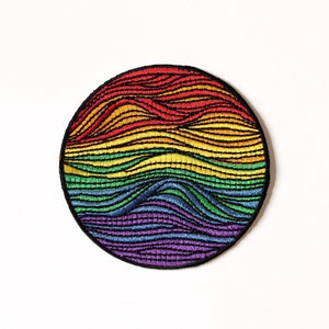 Può includere: Un cerotto rotondo ricamato con i colori dell'arcobaleno con un motivo ondulato. Il cerotto è in tessuto e ha un contorno nero.