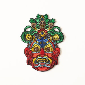 May include: Embroidered patch featuring a colorful, detailed mask with a fierce expression. The mask has a red face with yellow eyes, a blue nose, and a green and yellow crown with skulls.