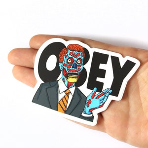 May include: A sticker featuring a cartoon character with a red and blue face, wearing a suit and tie, with the word "OBEY" in black letters.