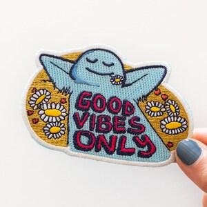 K&ouml;nnte beinhalten: Blauer gestickter Aufn&auml;her mit einer Comicfigur mit erhobenen Armen und dem Text "Good Vibes Only", umgeben von gelben G&auml;nsebl&uuml;mchen und roten Herzen.