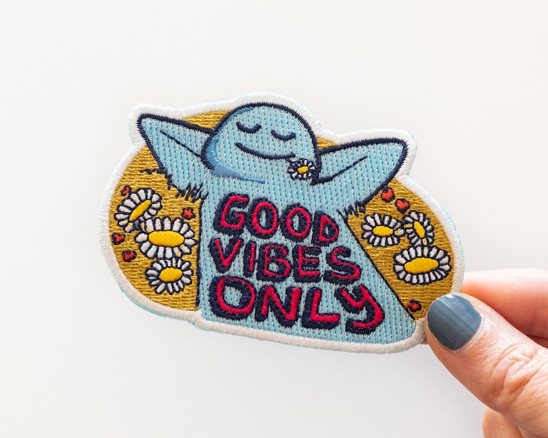 K&ouml;nnte beinhalten: Blauer gestickter Aufn&auml;her mit einer Comicfigur mit erhobenen Armen und dem Text "Good Vibes Only", umgeben von gelben G&auml;nsebl&uuml;mchen und roten Herzen.