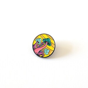 May include: A round enamel pin with a yellow and black striped background. The pin features a cluster of colorful mushrooms with pink, purple, and blue stems and green and blue caps.