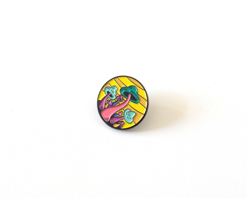 May include: A round enamel pin with a yellow and black striped background. The pin features a cluster of colorful mushrooms with pink, purple, and blue stems and green and blue caps.