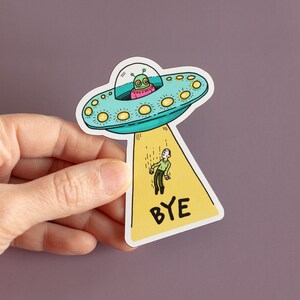 May include: A yellow and blue sticker with a cartoon alien spaceship and a person falling from it. The sticker says "BYE" in black letters.