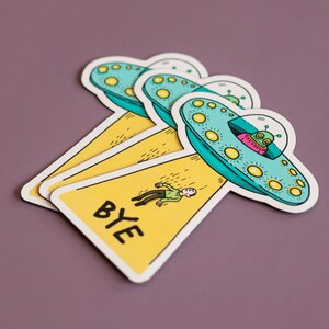 May include: Three yellow and blue alien spaceship stickers with a person falling from the top one. The bottom sticker says "BYE" in black letters.