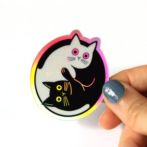 May include: A holographic sticker featuring a yin yang design with two cats. The white cat is on the left side of the sticker and the black cat is on the right side. The cats are facing each other and their tails are intertwined.