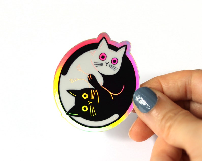 May include: A holographic sticker featuring a yin yang design with two cats. The white cat is on the left side of the sticker and the black cat is on the right side. The cats are facing each other and their tails are intertwined.