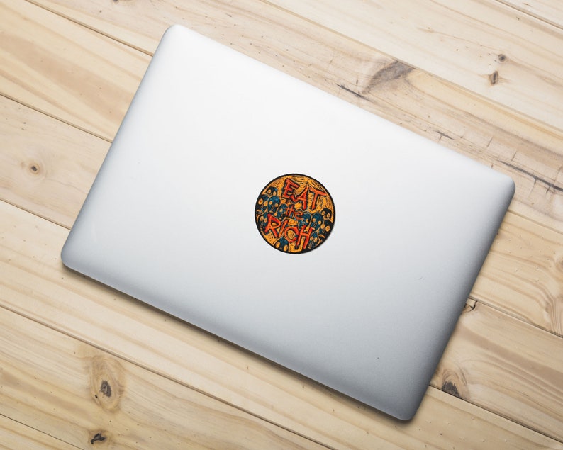 May include: A round sticker with a yellow background and a black outline. The sticker features a graphic of a crowd of people with their faces obscured by black circles. The text "EAT THE RICH" is written in red and orange letters.