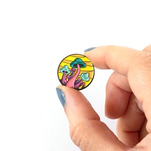 May include: A yellow and green enamel pin featuring a cluster of mushrooms with pink stems and blue caps against a yellow background.