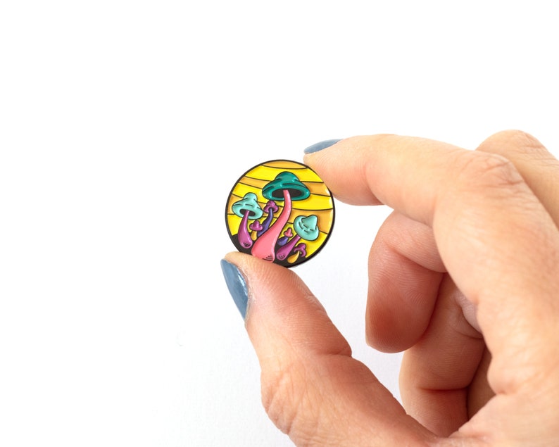 May include: A yellow and green enamel pin featuring a cluster of mushrooms with pink stems and blue caps against a yellow background.